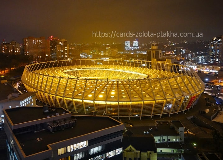 Aerial night view of Kyiv's Olimpiyskiy Stadium glowing gold amid the city lights.
