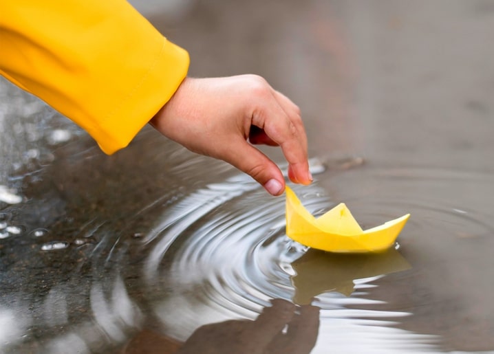 Child's hand releasing a yellow paper boat onto a rain puddle