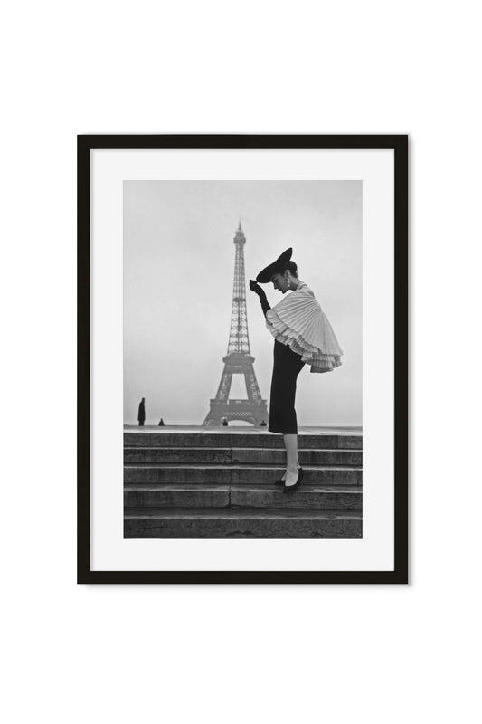 Black-and-white photo of a woman in a pleated cape and hat on steps, Eiffel Tower in the background, framed poster