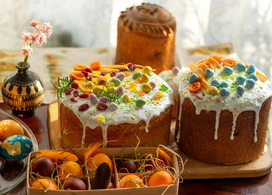 Two iced paska cakes with rosemary and pastel eggs beside a straw-filled egg box