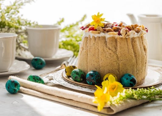 Frosted paska topped with walnuts, cranberries and a daffodil, surrounded by turquoise speckled eggs and tea cups.