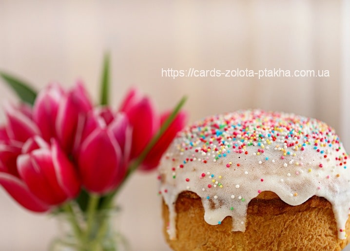Runde Osterpaska mit weißem Zuckerguss und bunten Streuseln neben rosa Tulpen