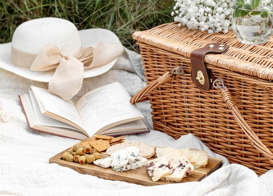 Picknickkorb, Käsebrett mit Oliven, Buch und Strohhut auf weißer Decke im Gras