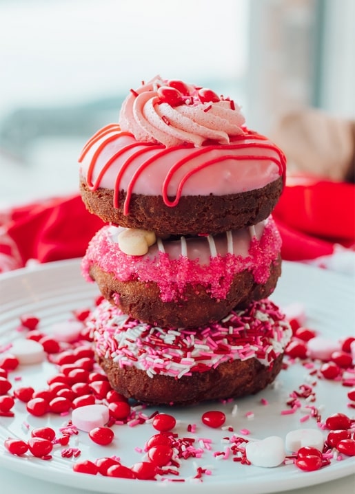 Three donuts with pink icing stacked on a plate, topped with sprinkles and candy hearts