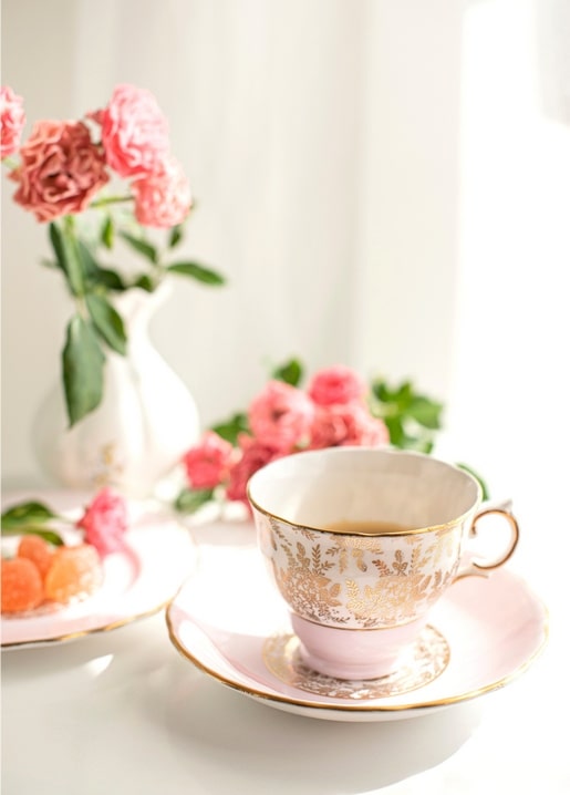 Gold-rimmed pink teacup with tea beside pink roses in a white vase