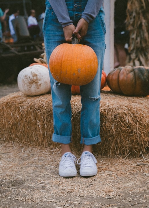 Cropped view of rolled jeans and white sneakers holding a pumpkin at a hay-covered pumpkin patch.