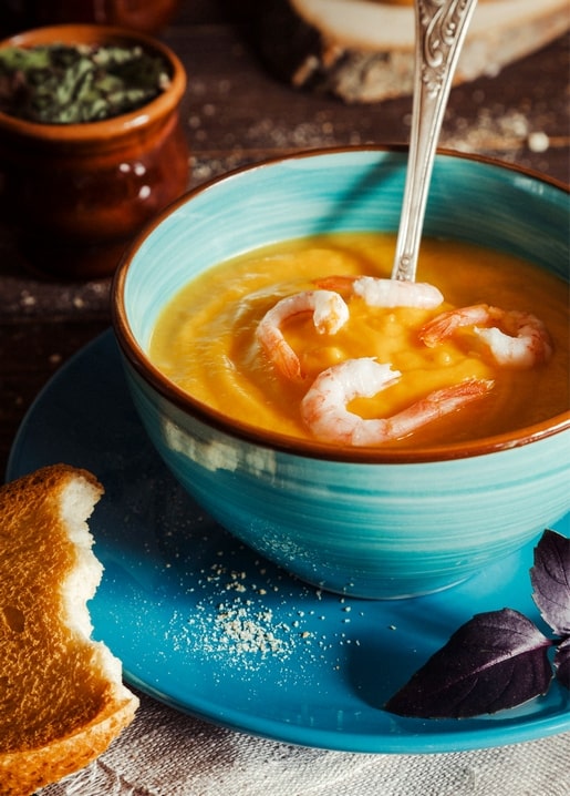 Bowl of pumpkin soup topped with shrimp on a teal plate with toasted bread and dried herbs.