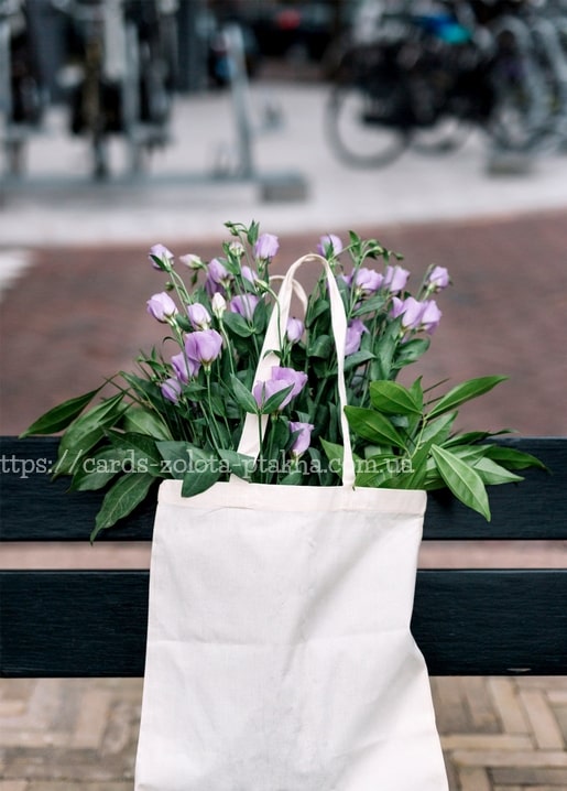 Lila Eustoma-Blüten quellen aus einer Stofftasche an einem schwarzen Geländer, im Hintergrund verschwommene Fahrräder