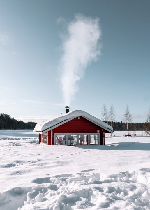 Rote Holzhütte am zugefrorenen See, Rauch steigt aus dem Schornstein in den blauen Winterhimmel.