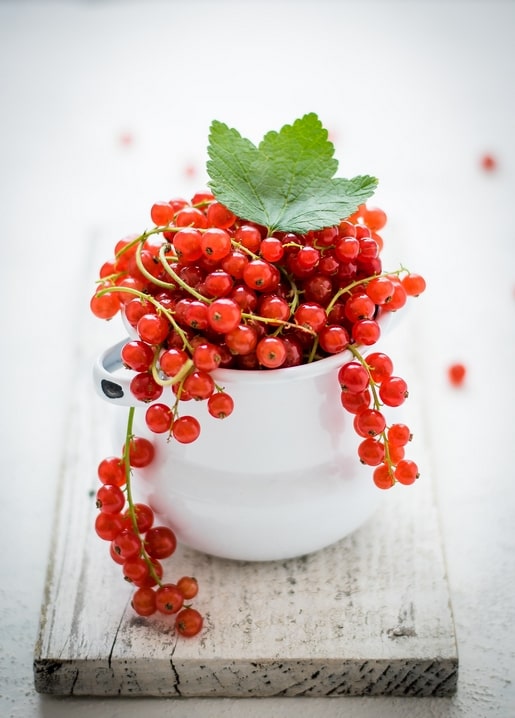 Reife rote Johannisbeeren quellen aus einem weißen Emaillebecher auf einem Holzbrett