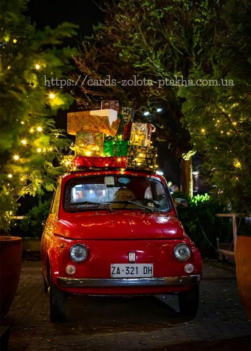 Roter Oldtimer-Fiat bei Nacht mit Weihnachtsgeschenken auf dem Dach zwischen beleuchteten Bäumen