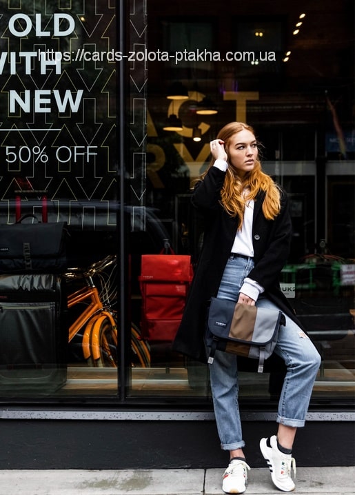 Rothaarige Frau im schwarzen Mantel lehnt an einem Schaufenster, Jeans und weiße Sneaker.