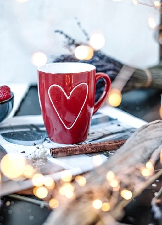 Rote Tasse mit weißem Herz, Zimtstange und Lichterkette auf einem gemütlichen Wintertisch