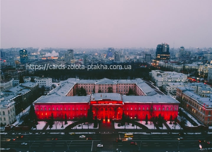 Aerial view of a large historic building lit red at winter dusk with snow and Kyiv skyline behind