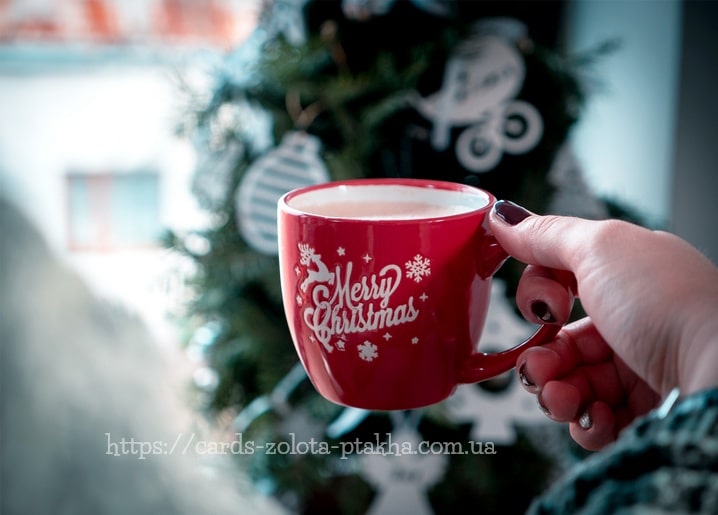 Hand hält rote Tasse mit „Merry Christmas“-Schriftzug und heißem Kakao vor Weihnachtsbaum