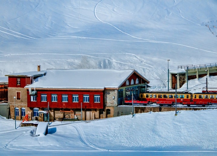 Red mountain train at a snow-covered alpine station with red buildings and white slopes