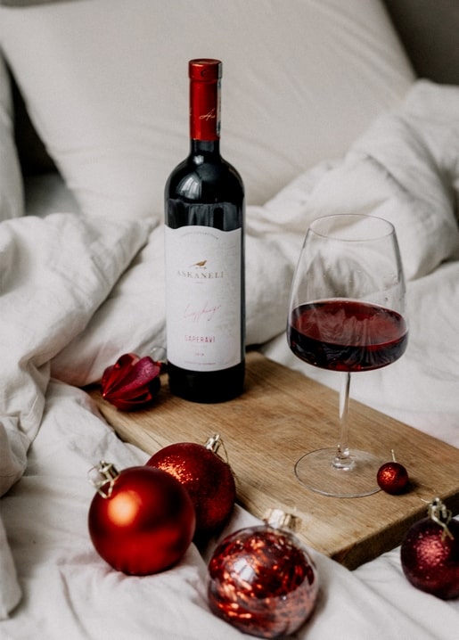 Bottle of red wine and a filled glass on a wooden tray among white bedding and red Christmas ornaments