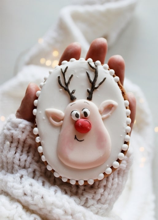 Gingerbread cookie iced as a smiling reindeer, held in a knit mitten by fairy lights