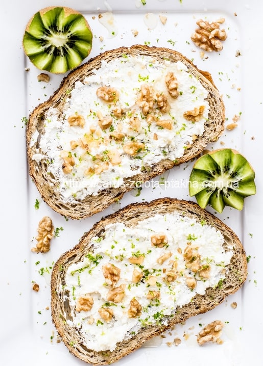 Ricotta-Toast mit Walnüssen und Limettenzeste neben frischen Kiwihälften auf weißem Teller