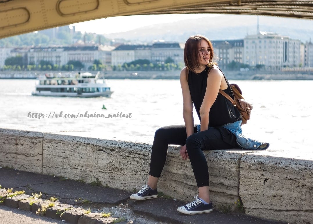 Young woman sitting on a stone riverside wall under a yellow bridge, boat passing on the river