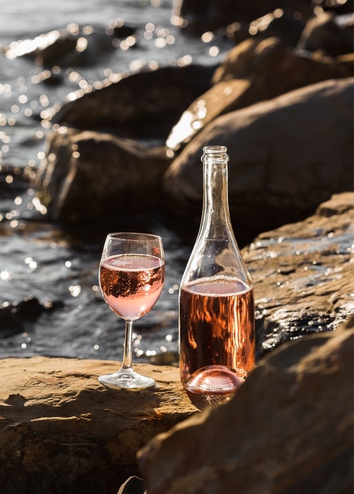 Flasche und Glas Roséwein auf Felsen am Meer bei Sonnenuntergang
