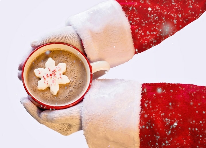 Two hands in Santa's red-and-white gloves holding a mug of cappuccino topped with a snowflake marshmallow