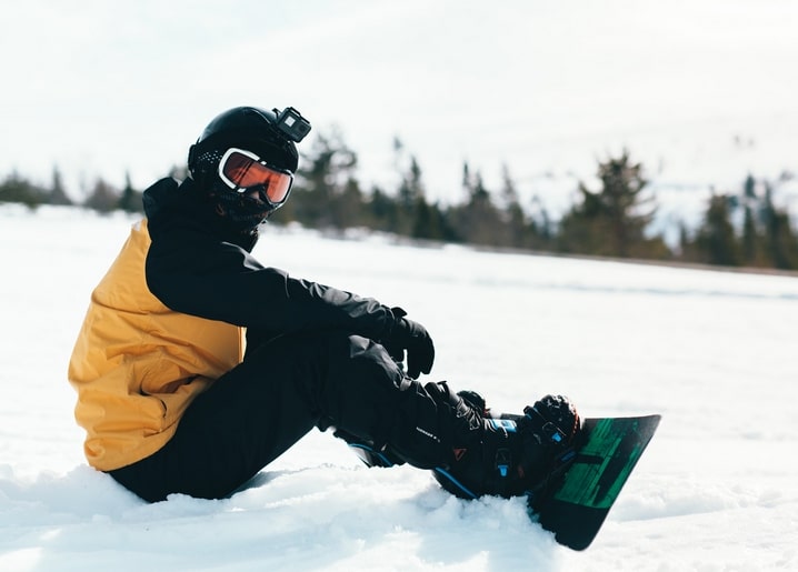 Snowboarder sitzt im Schnee in gelber Jacke mit Helmkamera, im Hintergrund ein Kiefernwald.