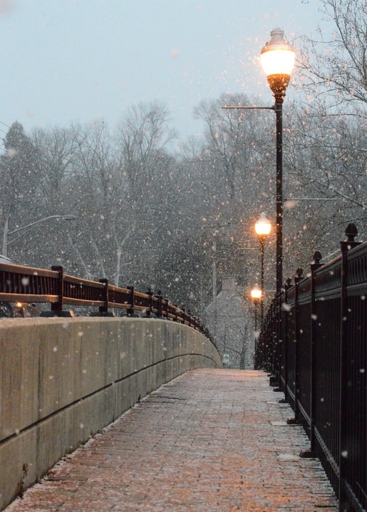 Row of glowing lamp posts along a snow-dusted stone bridge walkway at dusk