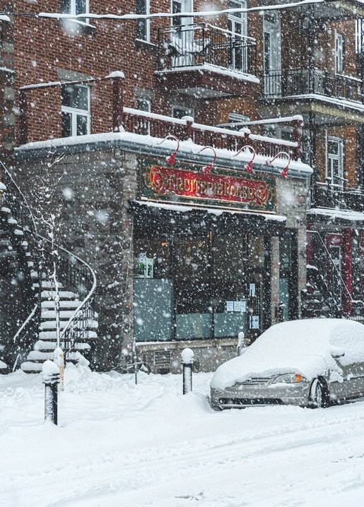 Schneebedecktes Auto vor einem beleuchteten Café-Schild in einer verschneiten Straße