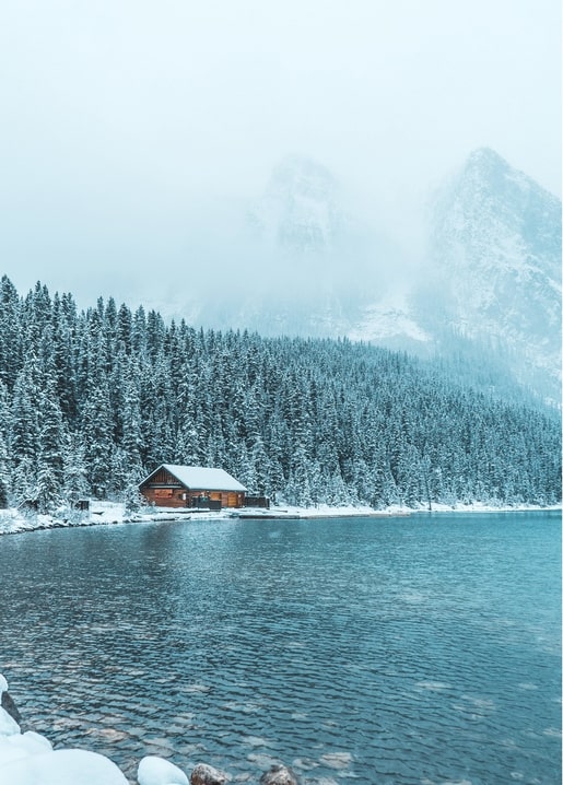 Snow-covered wooden cabin by a lake with foggy mountains behind