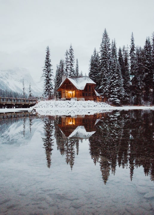 Verschneite Blockhütte mit erleuchteten Fenstern spiegelt sich im Bergsee bei Dämmerung