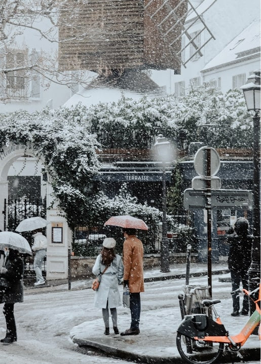 Paar mit Schirm auf einer verschneiten Pariser Straße vor einer alten Windmühle