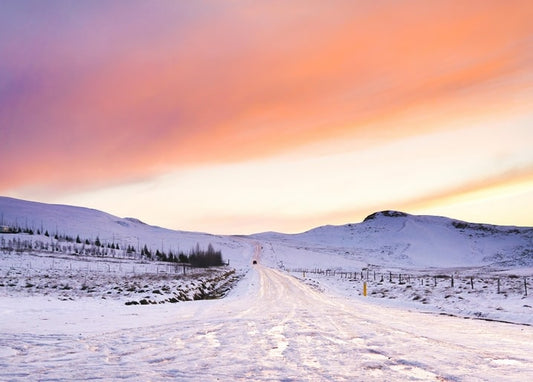 Snow-covered road leading toward mountains under a pink sunset sky
