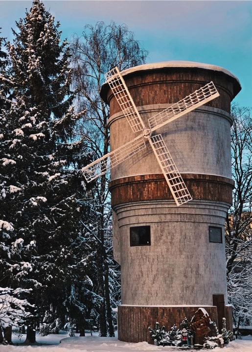 Schneebedeckte Holzwindmühle mit weißen Flügeln neben verschneiten Kiefern in der Dämmerung