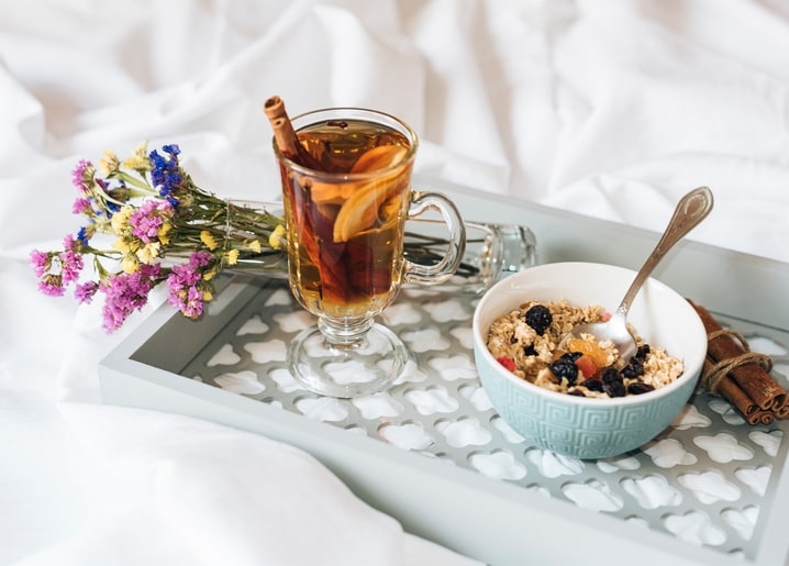 Glass mug of spiced tea, a bowl of berry granola, and wildflowers on a white breakfast tray in bed