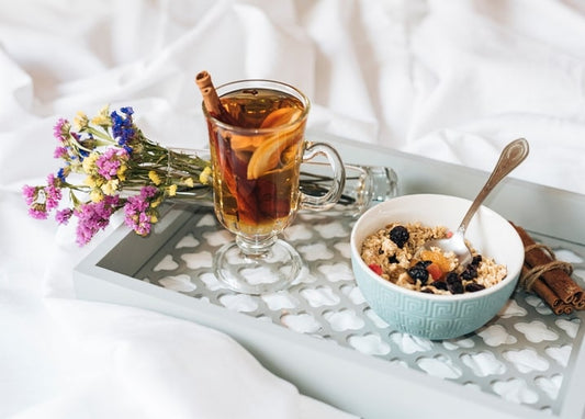 Glass mug of spiced tea, a bowl of berry granola, and wildflowers on a white breakfast tray in bed