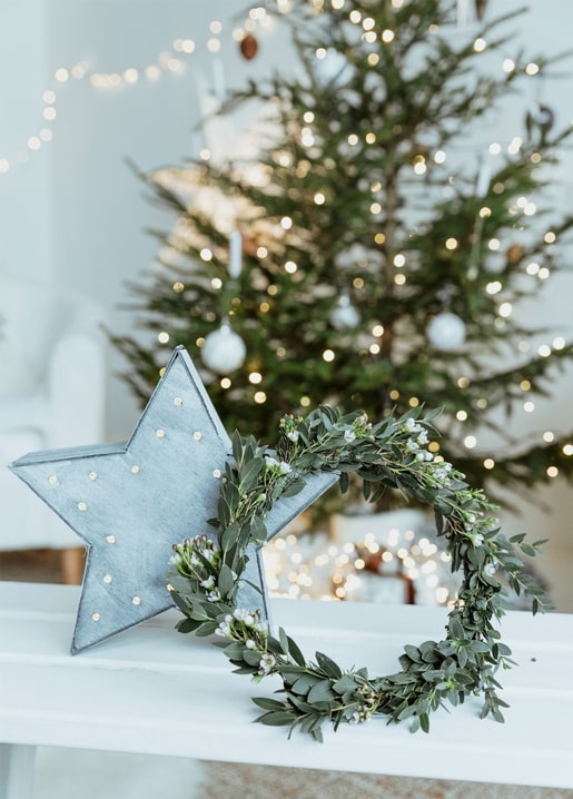 Metallener Sternschmuck neben einem Eukalyptus- und Winterbeerenkranz auf einer weißen Bank vor einem beleuchteten Weihnachtsbaum