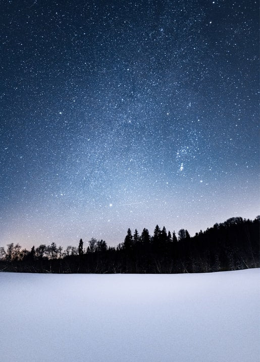 Countless stars and the Milky Way over a snow-covered field and dark tree line at night