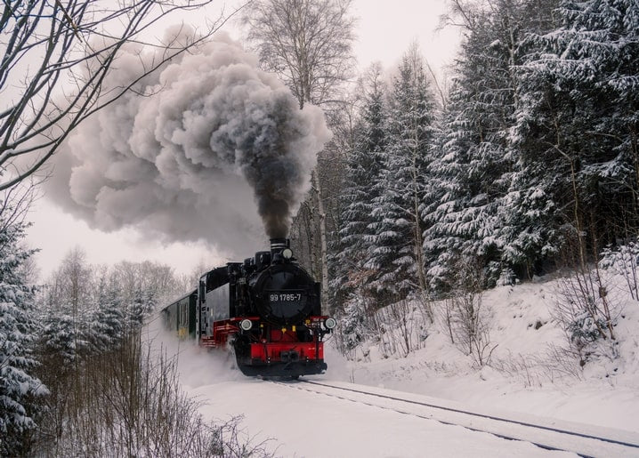 Red steam locomotive pulling train cars through a snowy pine forest, billowing smoke against the sky.