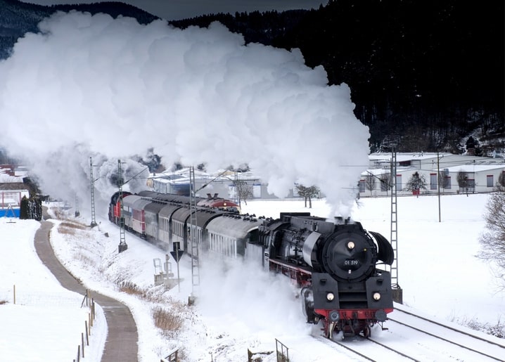Black steam locomotive pulling carriages through a snowy mountain valley with billowing smoke