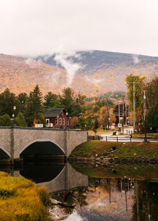 Stone arch bridge reflecting in a river beneath misty, autumn-colored mountains near a small village.