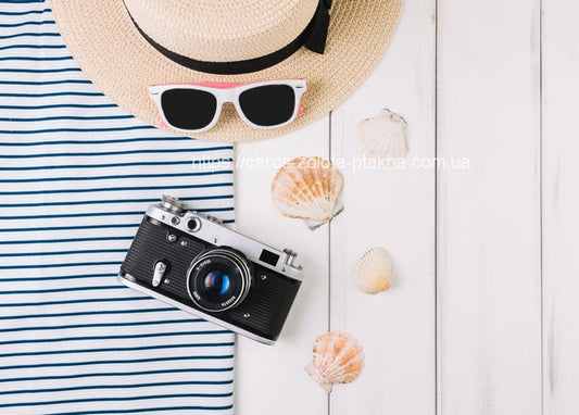 Flatlay mit Strohhut, gestreiftem Shirt, Sonnenbrille, Retro-Kamera und Muscheln auf weißem Holz
