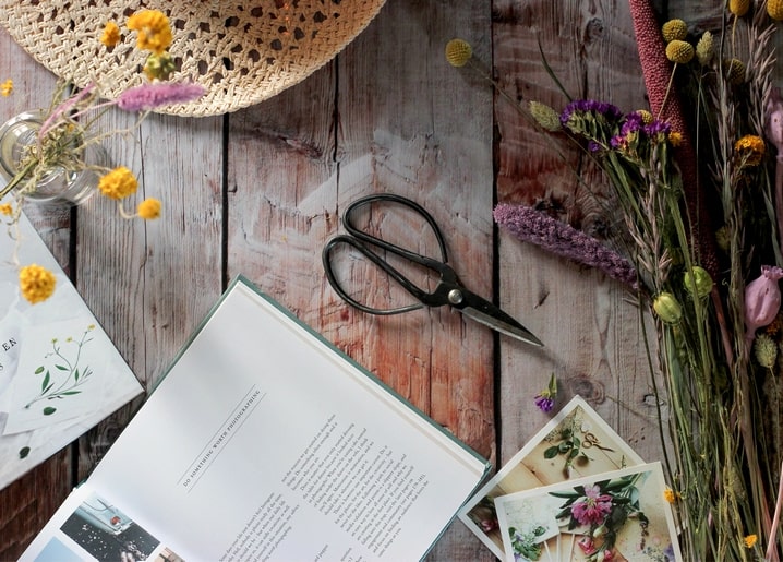 Strohhut, Schere und getrocknete Blumen auf einem Holztisch neben Postkarten