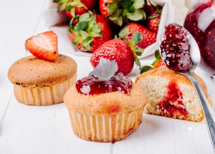 Aufgeschnittener Cupcake mit Erdbeermarmelade-Füllung, frischen Erdbeeren und einem Löffel Marmelade