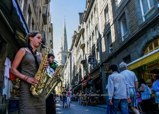 Zwei Straßenmusiker spielen Saxophon in einer Kopfsteinpflastergasse, im Hintergrund ein Kirchturm