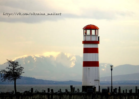 Rot-weißer Leuchtturm vor schneebedeckten Bergen und ruhigem Wasser in der Abenddämmerung