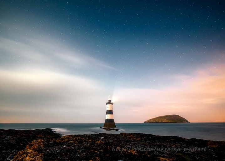 Schwarz-weiß gestreifter Leuchtturm mit Lichtstrahl unter Sternenhimmel neben kleiner Insel