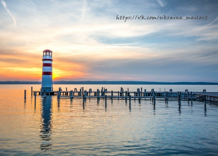 Rot-weiß gestreifter Leuchtturm vor Sonnenuntergang neben einem langen Holzsteg über ruhigem Wasser