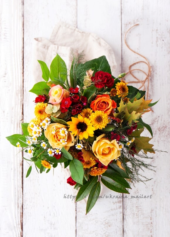 Strauß aus Sonnenblumen, goldenen Rosen und weißen Gänseblümchen, in Leinen auf weißem Holz gebunden.