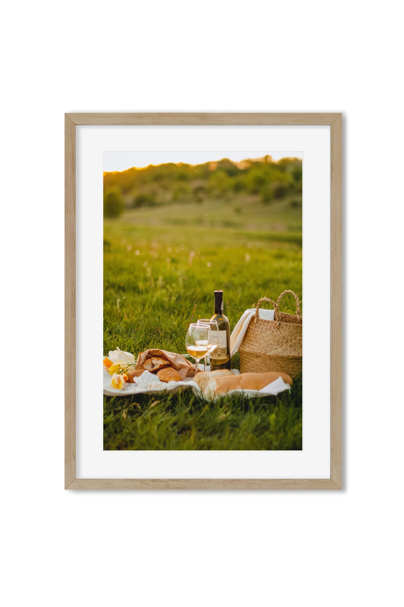 Golden-hour picnic scene with wine, bread, flowers and a wicker basket in a green meadow